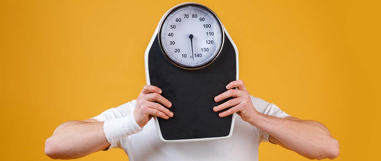 Young sports man hiding face behind weight scales